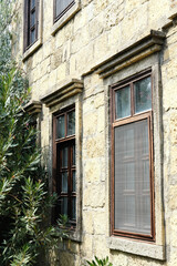 Fototapeta na wymiar Stone Building with Windows and Lush Greenery. Tall windows with dark frames set in a textured stone wall, partially obscured by lush green oleander shrubs. Vertical photo