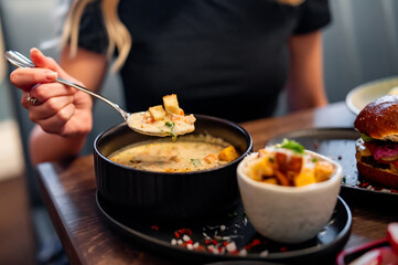 “Person enjoying a creamy soup, served with a burger and fries in a cozy indoor setting. Focus on the food and hand holding the spoon - obrazy, fototapety, plakaty
