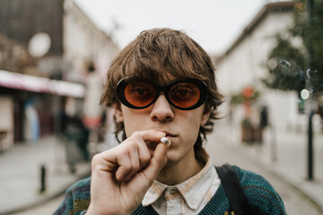 Fashionable young man in retro 90s style attire smokes cigarette on bustling city street, exuding...