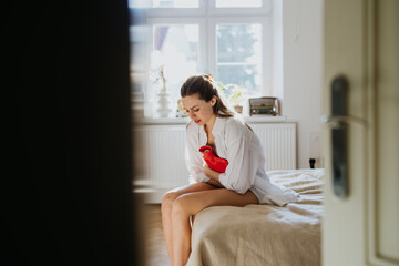 Woman at home suffering from menstrual pain, having cramps. Woman warming lower abdomen with a hot...