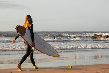 Female surfer dressed in wetsuit holding her surfboard walking along the beach shore