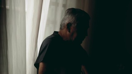 Pensive senior man standing in the shadows indoors during challenging times. Depressed thoughtful person struggles with old age and illness