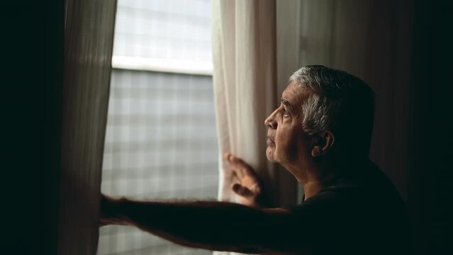 Thoughtful Senior Man Peering Through Window Curtains, Elderly Observing Neighborhood from Inside Home