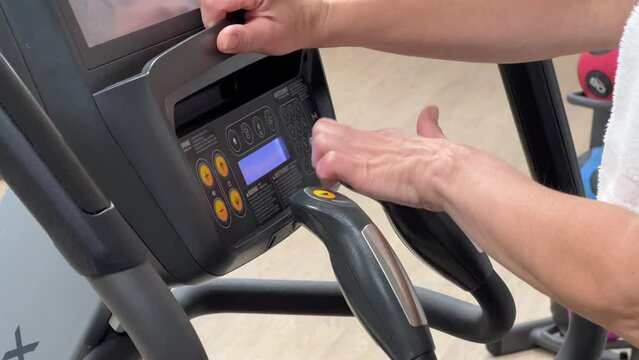 An elliptical cross-trainer is programmed for cardio training by a woman's hand. Hands of an elderly woman on the exercise machine. Horizontal video of orbitrek setup on a blurred background of the