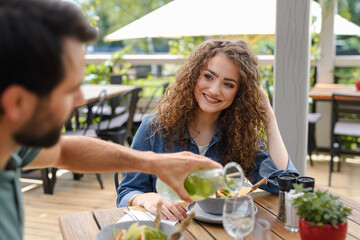 Young couple at date in restaurant, sitting on restaurant terrace. Boyfriend pouring water in girlfriends glass, taking care of her. - 755173595