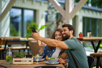 Young couple at date in restaurant, taking selfie, sitting on restaurant terrace. Boyfriend and girlfriend enjoying springtime, having lunch outdoors. - 755173965