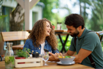 Young couple laughing at date in restaurant, sitting on restaurant terrace. Boyfriend and girlfriend enjoying springtime, having lunch outdoors. - 755174193