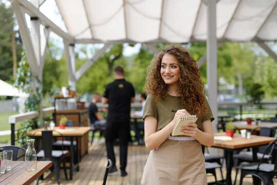 Portrait of a beautiful waitress writing down an order in paper notebook with pen. Server standing on restaurant terrace in an apron.