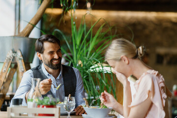 Happy loving couple at date in restaurant. Husband and wife sitting at table, laughing, having lunch in modern cafe. - obrazy, fototapety, plakaty