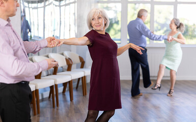 Happy elderly lady with man in elegant clothes dancing bachata in modern ballroom dance class