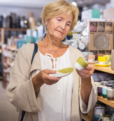 Elderly female customer chooses plates and cups in tableware store