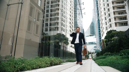 Low angle camera view of caucasian businessman calling to manager and hold suitcase while walking at skyscraper view. Skilled project manager working on telephone and sharing marketing ideas. Urbane.