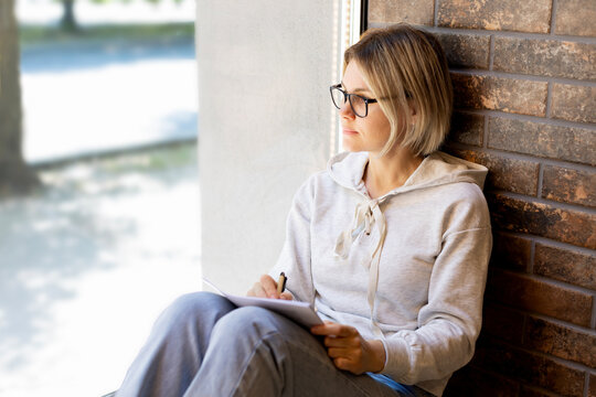 Blonde woman in glasses looking thoughtfully out the window and writing in a notebook. A student is studying, doing homework, or writing a book. Serious girl in grey sweater, sunny summer day