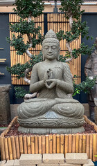 Stone Buddha figure sitting in a indoor garden with a background of oriental trees and bamboo