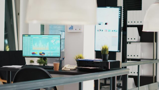 Office with computer monitor on desk showing stock exchange listings and display in background with business charts. PC in workspace with trading platform indexes and company data graphs in the back
