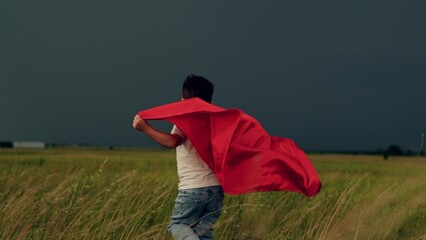 Little boy in red mantle play superhero against gray overcast sky. Happy kid run field in red...