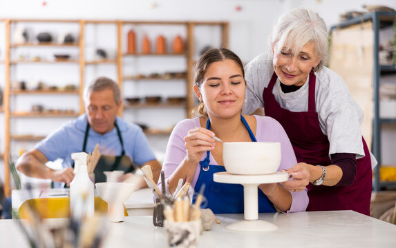 Young woman in class with elderly woman potter to learn how to paint clay vase