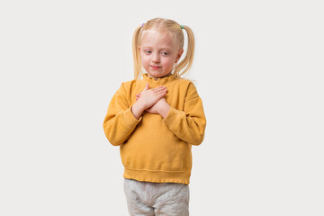 Adorable Caucasian little girl with blond hair in a yellow hoodie standing with folded hands on a white background