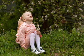 Little blond girl sitting on the ground and looking up at the sky. Child in blooming garden. Summer day