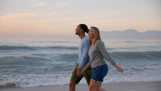 Young couple wearing casual clothing holding hands and running along beach through surf at dawn - shot in slow motion