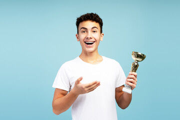 Excited boy with braces wearing white t shirt, holding trophy cup, pointing at it, looking at camera