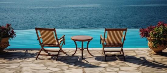 Fototapeta premium A pair of chairs are positioned next to a table on the deck of a swimming pool, overlooking the clear blue water of a hotel resort on a sunny summer day.
