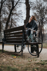Stylish young businesswoman takes a thoughtful break on a park bench alongside her bicycle, emanating elegance and confidence.