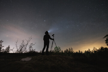 Male landscape astrophotographer with a camera on a tripod outdoors in early spring at night under the starry sky.