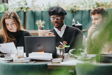 Multicultural group of creative people collaborates on a project, working intently at a laptop in a relaxed restaurant setting surrounded by greenery.