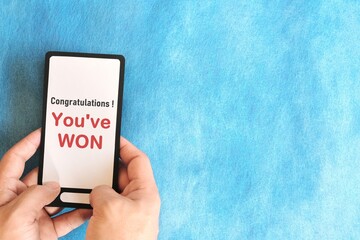 Winning text message scam concept. Hand holding mobile phone with congratulatory message in blue background.