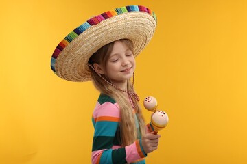 Cute girl in Mexican sombrero hat dancing with maracas on orange background