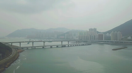 Obraz premium Aerial view of a curving bridge over water with urban skyscrapers against a backdrop of hazy mountains