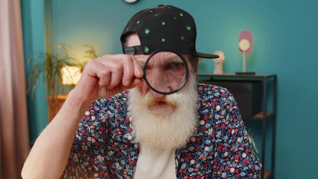 Investigator researcher scientist senior man working at home office holding magnifying glass near face looking into camera with big zoomed funny eye, searching, analyzing. Amazed stylish grandpa