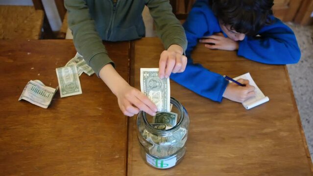 Siblings aged 6 and 8 keep dollar bills and save money in a glass jar. Concept of financial education, saving, investing with children and economy.