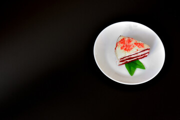 White ceramic plate with a slice of red velvet cake with mint leaves and sprinkles on a black background.