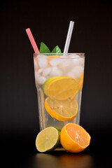 A tall glass of refreshing lemonade with ice and citrus fruit pieces on a black background, next to ripe lime and orange slices.