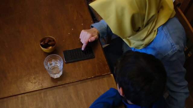 Mother with son reading the quran on the mobile phone in front of some dates and a glass of water in ramadan eid fitar i religious spiritual education islam muslim with hijab