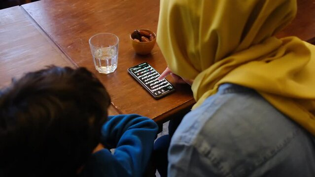 Mother with son reading the quran on the mobile phone in front of some dates and a glass of water in ramadan eid fitar i religious spiritual education islam muslim with hijab