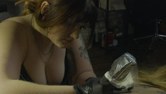 a female tattoo artist  wearing black rubber gloves works intently on tattoing her clients back