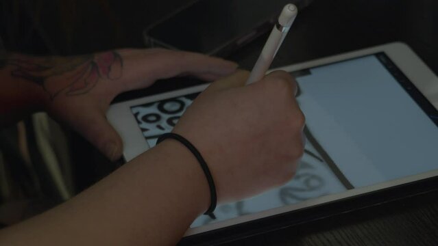 close up shot of a woman's tattooed hands as she  uses a stylus to draw a design on an electronic tablet