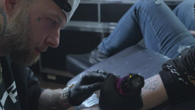 a white bearded male tattoo artist uses an iron or tattoo pen to draw a tattoo onto his female clients upper arm