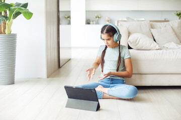 Head shot happy cute little 9s old kid girl wearing earphones, looking in digital tablet screen at home