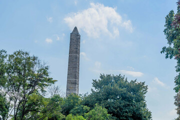Historic stone obelisk rising beyond tree tops against a blue sky, in Istanbul, Turkiye