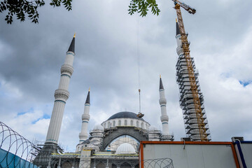 Mosque minarets rise into cloudy skies, flanked by construction equipment and scaffolding,