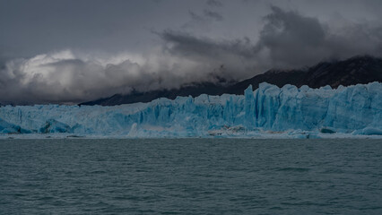 A wall of blue cracked ice stretches over a glacial lake. Melted ice floes float in turquoise...
