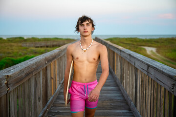 Handsome teenage male model carrying his skim board while walking from the beach on a long wooden bridge  - Powered by Adobe