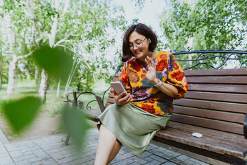 Cheerful adult woman with glasses engaged in a lively phone conversation while seated in green park. Joyful female with smartwatch, enthusiastically conversing on her phone on a sunny day in park