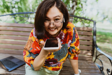 Happy woman sitting on a bench in a green park, emotionally talking on her phone. Smiling freelancer wearing a colorful blouse, animatedly chatting on her smartphone outdoor.