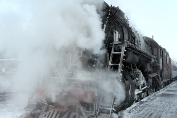 A steam locomotive, wrapped in steam, stands on the platform on a frosty winter day. A locomotive with ice-covered parts on the hull.