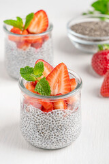 Organic healthy sweet chia pudding dessert made with seeds and plant based milk decorated with sliced raw ripe strawberries topping and fresh mint leaf served in glass jar on white wooden table